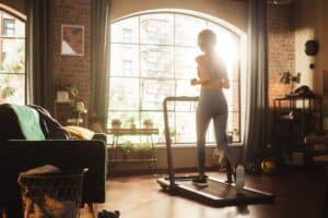 Beautiful Athletic Sports Woman Running on a Treadmill at Her Home Gym. Workout Female Athlete Training while Listening Podcast or Music in Headphones. Apartment with Window View. Side Back View